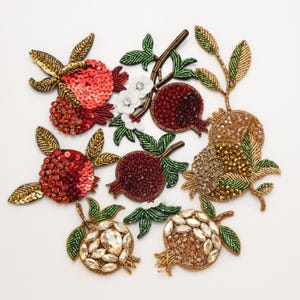 May include: A collection of pomegranate-shaped patches featuring intricate beadwork, sequins, and crystals in shades of red, gold, and green. Each patch displays detailed leaves and stems, with some designs incorporating white floral accents and metallic thread.