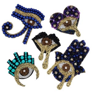 Può includere: Cinque toppe ricamate con un designo di occhio malvagio. Le toppe hanno la forma di un cuore, una mano e un ankh. Le toppe sono decorate con perline blu, viola, turchese e dorate.