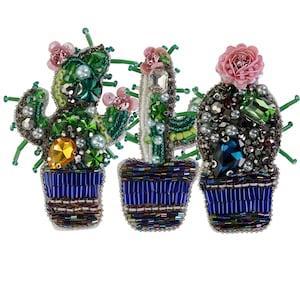 Può includere: Tre piante di cactus perlate in vasi blu. Ogni cactus è decorato con perline colorate, strass e perle. I cactus sono disposti in fila da sinistra a destra.
