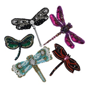 May include: Five decorative dragonfly brooches in various colors. The brooches are embellished with beads, sequins, and pearls. Colors include black, teal, green, red, and purple. The brooches are likely for clothing or accessories.