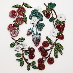 May include: A collection of five embroidered patches featuring fruit and floral motifs. The designs include beaded red berries, strawberries, and white flowers with green leaves, all crafted with intricate beadwork and sequins on fabric.
