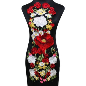Op de afbeelding: Een zwarte jurk met een groot geborduurd bloemenmotief. Het motief bevat rode, witte en gele bloemen met groene bladeren.