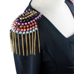 Puede incluir: Un blazer negro con una pieza decorativa en el hombro. La pieza del hombro presenta filas de tachuelas puntiagudas de colores en oro, rosa, morado y azul, con cadenas doradas colgantes. El blazer se exhibe en un maniquí.