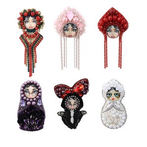 May include: A collection of six decorative brooches featuring painted faces and elaborate beadwork. The brooches showcase various designs, including a Matryoshka doll, a bow, and headpieces adorned with beads and crystals in red, pink, and white.