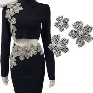 Könnte beinhalten: Ein schwarzes Kleid mit einem Strass-Blumenmuster auf dem Oberteil und dem Rock. Das Kleid hat eine figurbetonte Silhouette und lange Ärmel. Drei zusätzliche Strass-Blumen werden separat gezeigt.