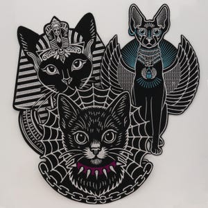May include: Three embroidered patches featuring black cats. One cat wears an Egyptian pharaoh headdress, another is a winged cat with a blue scarab amulet, and the third is a cat framed by a spider web with a purple spiked collar and chain.