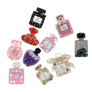 May include: A collection of decorative perfume bottle-shaped embellishments. The bottles are adorned with beads, gems, and bows in various colors, including pink, red, black, and purple. Each bottle features a label with the text "N°5 PARIS".
