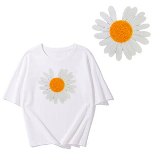 Puede incluir: Camiseta blanca con una margarita blanca bordada en la parte delantera. La margarita tiene un centro amarillo.