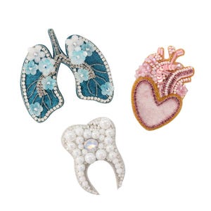 May include: Three handmade brooches: a teal lung design with white and blue floral accents, a pink heart with beaded details, and a white tooth with pearl embellishments. Each brooch is intricately crafted with detailed stitching.