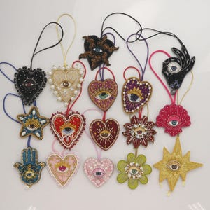 May include: A collection of fourteen decorative ornaments featuring beaded designs of hearts, stars, flowers, shells, and hands, each centered with an eye motif. The ornaments are crafted with colorful beads, sequins, and pearls, and include attached hanging loops.