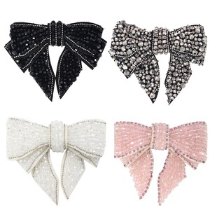Peut inclure: Quatre décorations en forme de nœud en strass. Les nœuds sont en noir, argent, blanc et rose.