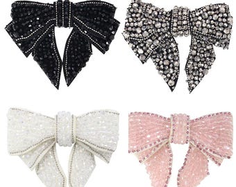 Puede incluir: Cuatro adornos en forma de lazo hechos de pedrería. Los lazos son de color negro, plateado, blanco y rosa.
