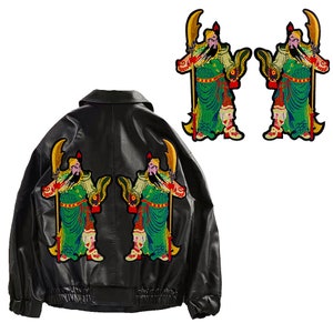 Pode incluir: Jaqueta de couro preta com dois patches bordados de uma figura de guerreiro segurando uma lança. O guerreiro está vestindo uma túnica verde e vermelha com detalhes dourados.