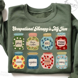 May include: A military green sweatshirt with the text "Occupational Therapy is My Jam" printed on the front. The text is in a retro font. There are nine jars of jam with different labels on the front of the sweatshirt. The labels include "play", "fine motor", "ADLS", "visual motor", "learn", "cognition", "social", and "sensory".