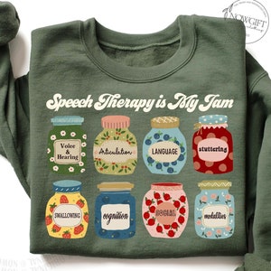 May include: A green sweatshirt with the text "Speech Therapy is My Jam" in a retro font. The text is surrounded by illustrations of jars of jam, each labeled with a different aspect of speech therapy, such as "Voice & Hearing", "Articulation", "Language", "Stuttering", "Swallowing", "Cognition", "Social", and "Medalities".