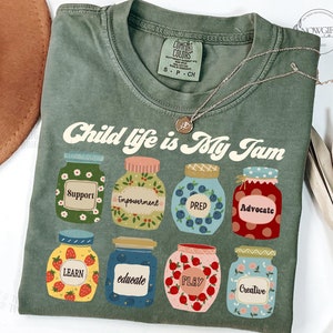 May include: A green t-shirt with the text "Child life is My Jam" in a retro font. The shirt features a design of eight jars with different words on them, including "Support", "Empowerment", "Prep", "Advocate", "Learn", "Educate", "Play", and "Creative".