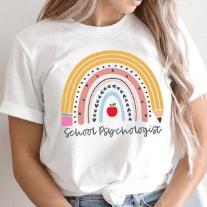 May include: White t-shirt with a rainbow design featuring a red apple and pencils. The text "School Psychologist" is printed below the rainbow.