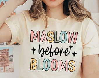 May include: A woman wearing a light beige t-shirt with the text "MASLOW +before+ BLOOMS" in colorful letters. The shirt has a relaxed fit and short sleeves. She is wearing a brown hat and blue jeans.