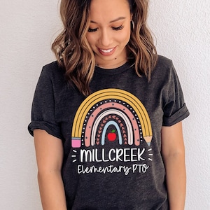 May include: A dark grey t-shirt featuring a rainbow design made of pencils and a red apple in the centre. The text "MILLCREEK Elementary PTO" is printed below the rainbow.