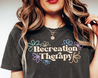 May include: A dark gray t-shirt with the text "Recreation Therapy" in a retro font with colorful flower accents.
