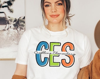 May include: White t-shirt with a colorful graphic of the letters "CES" with the text "Crestline Elementary" underneath.