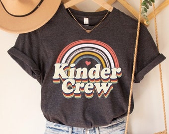 May include: A dark gray t-shirt with a rainbow and the text "Kinder Crew" in a retro font. The shirt is on a hanger with a straw bag and a hat.