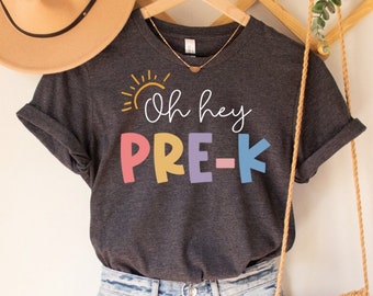 Może przedstawiać: Ciemnoszara koszulka z napisem "Oh hey PRE-K" w kolorowych literach. Koszulka jest połączona z dżinsowymi szortami, kapeluszem w kolorze beżowym i małą torebką z makramy z frędzlami. Widoczny jest również złoty naszyjnik.