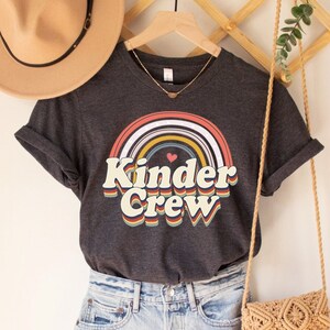 May include: A dark grey t-shirt with a rainbow and the text "Kinder Crew" in a retro font. The shirt is on a hanger with a straw bag and a hat.