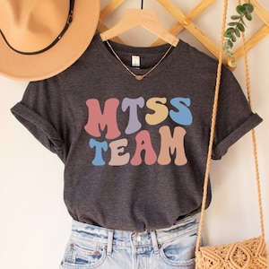 May include: A dark gray t-shirt with a colorful retro-style graphic that says "MTSS TEAM". The shirt is hanging on a wooden hanger with a straw hat and a woven bag.