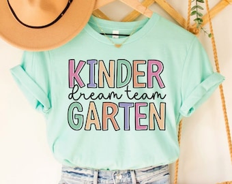 Może przedstawiać: Jasno błękitny t-shirt z napisem "kinder dream team garten" w kolorach tęczy. Koszulka wisi na drewnianym wieszaku z brązowym kapeluszem i brązową plecioną torbą.