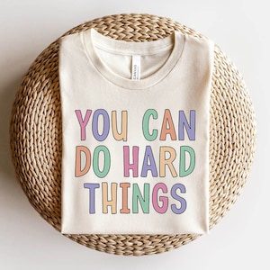 Peut inclure: Un t-shirt beige avec le texte "YOU CAN DO HARD THINGS" en lettres colorées. Les lettres sont soulignées de noir et ont un schéma de couleurs arc-en-ciel.