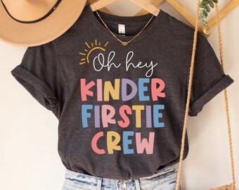 Może przedstawiać: Szary T-shirt z nadrukiem żółtego słońca i tekstem "Oh hey Kinder Firsttie Crew" w kolorowych literach. Koszulka wisi na drewnianym wieszaku z plecioną torbą i brązowym kapeluszem.