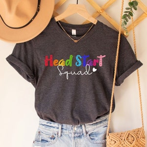 May include: A dark gray V-neck t-shirt with the text "Head Start Squad" in rainbow colors. The shirt is hanging on a wooden hanger with a straw bag and a hat.