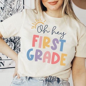 May include: A white t-shirt with a colorful graphic that says "Oh hey first grade" with a yellow sun graphic above the text.