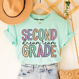 May include: A light blue t-shirt with the text "SECOND dream team GRADE" in colorful letters. The shirt is on a hanger with a straw hat and a woven bag.