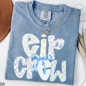May include: A blue jean Comfort Colors t-shirt with the words "Elf Crew" in a patchwork design. The letters are decorated with floral and gingham patterns, and a blue bow. A gold necklace with a pendant is draped across the shirt.