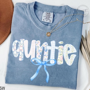 Könnte beinhalten: Ein blaues Comfort Colors T-Shirt mit dem Wort "Auntie" in einem Patchwork-Design. Die Buchstaben zeigen florale, gestreifte und Vichy-Muster. Eine blaue Schleife befindet sich unten, und eine goldene Halskette mit Anhänger ist auf dem Shirt.