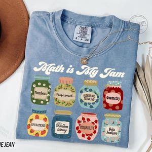 May include: A blue jean t-shirt with the text "Math is My Jam" printed on it. The shirt features a graphic of jars with different math-related terms, such as "Numbers", "Measurement", "Algebraic Thinking", "Geometry", "Operations", "Problem Solving", "Probability", and "Data Analysis".