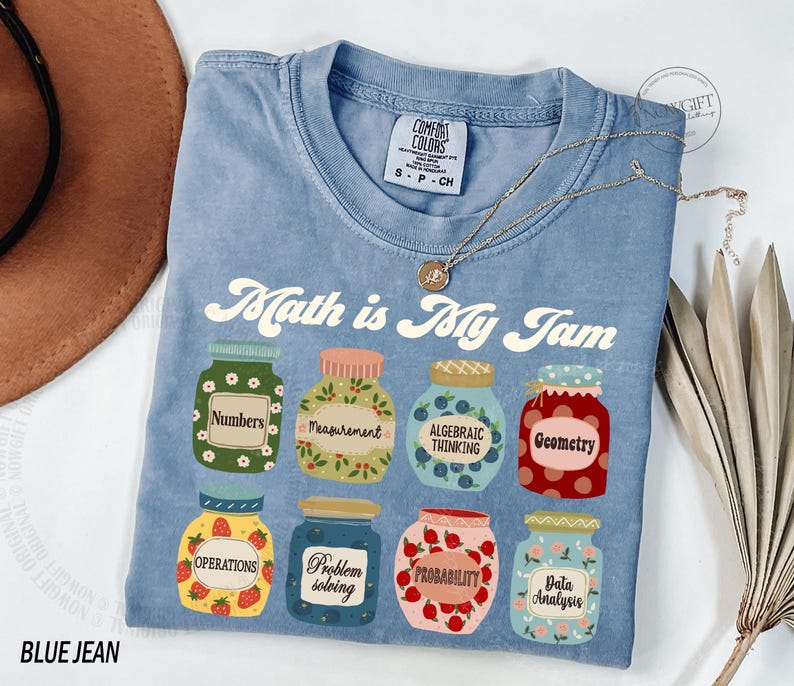 May include: A blue jean t-shirt with the text "Math is My Jam" printed on it. The shirt features a graphic of jars with different math-related terms, such as "Numbers", "Measurement", "Algebraic Thinking", "Geometry", "Operations", "Problem Solving", "Probability", and "Data Analysis".