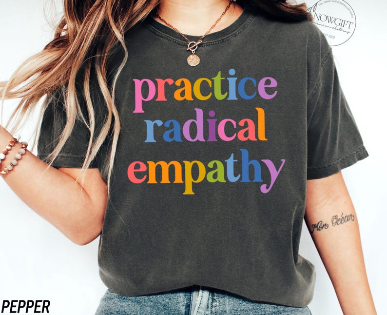 May include: Dark gray t-shirt with the words "practice radical empathy" in colorful, rainbow-colored letters. The shirt is a crew neck style and appears to be made of a soft, comfortable material. The word "pepper" is printed at the bottom left.