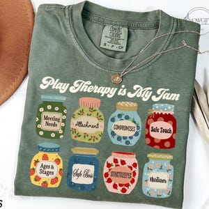 May include: A green t-shirt with the text "Play Therapy is My Jam" printed on the front. The text is in a retro font and is surrounded by illustrations of jars with different labels, such as "Meeting Needs", "Attachment", "Compromises", "Safe Touch", "Ages & Stages", "Safe Boss", "Storykeeper", and "Nurturer".