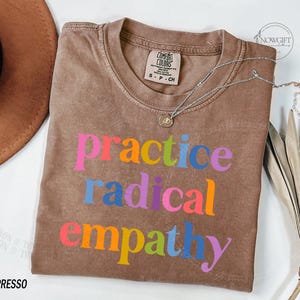 May include: Espresso-colored t-shirt with the words "practice radical empathy" in colorful block letters. The shirt has a crew neck and a Comfort Colors tag. A silver necklace with a small pendant is draped across the shirt.