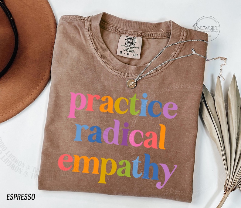 May include: Espresso-colored t-shirt with the words "practice radical empathy" in colorful block letters. The shirt has a crew neck and a Comfort Colors tag. A silver necklace with a small pendant is draped across the shirt.