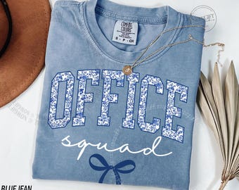 Op de afbeelding: Een blauw Comfort Colors T-shirt met de woorden "OFFICE squad" in witte letters en een blauwe strik. Het woord "OFFICE" is in een universiteitslettertype met een blauw bloemenpatroon. Een gouden ketting ligt op het shirt.