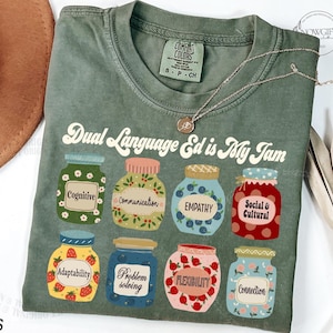 May include: Moss green t-shirt with the text "Dual Language Ed is My Jam." The shirt features illustrations of eight colorful jam jars, each labeled with a different concept, such as "Cognitive" and "Empathy." A delicate gold necklace rests on the shirt.