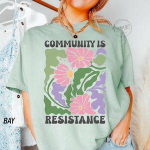 May include: A light green t-shirt with a graphic print of stylized flowers and leaves. The text "COMMUNITY IS RESISTANCE" is printed in black letters.