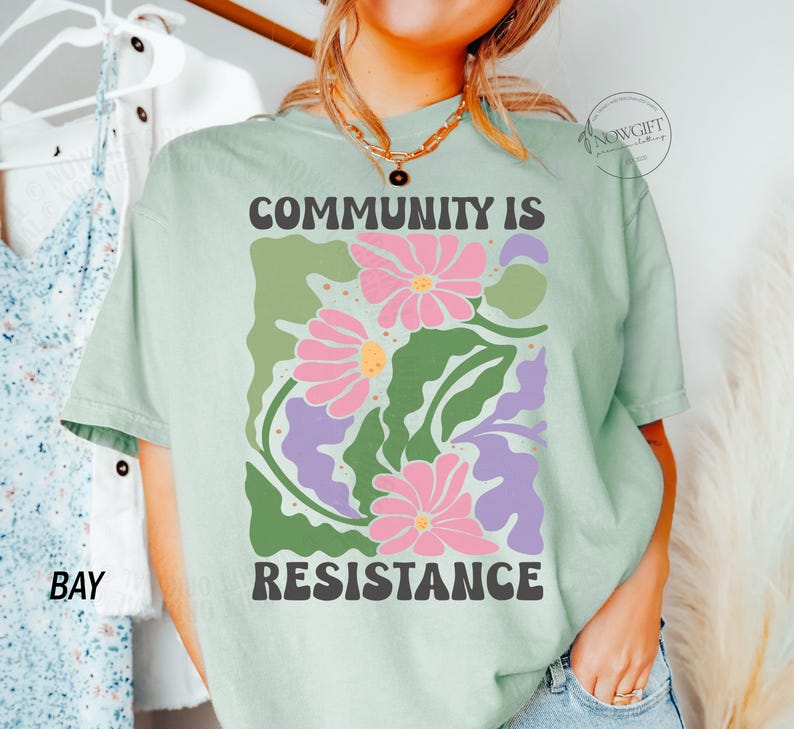 May include: A light green t-shirt with a graphic print of stylized flowers and leaves. The text "COMMUNITY IS RESISTANCE" is printed in black letters.
