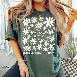 May include: Moss green t-shirt with a daisy design and the text "Helping You Grow To Your Full Potential Occupational Therapy". The shirt has a crew neck and short sleeves.