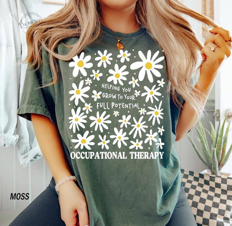 May include: Moss green t-shirt with a daisy design and the text "Helping You Grow To Your Full Potential Occupational Therapy". The shirt has a crew neck and short sleeves.