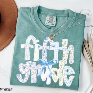 May include: Light green t-shirt with the words "Fifth Grade" in a patchwork design. The letters are decorated with floral and gingham patterns, and a blue bow. A gold necklace is draped across the shirt.